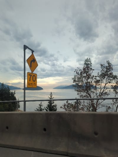 The image displays a static outdoor scene captured from a roadside perspective overlooking a large body of water, consistent with a coastal highway segment within Electoral Area A, British Columbia, Canada, characteristic of the Sea-to-Sky corridor.
In the foreground, a dark gray concrete barrier extends horizontally across the lower third of the frame, exhibiting two circular depressions. Above this barrier, a metallic gray guardrail with two parallel horizontal bars is visible. To the left, a dark gray pole supports two stacked, yellow, diamond-shaped road signs. The upper sign features a black curved arrow indicating a sharp right turn. The lower sign displays "70 km/h" in black text, denoting a speed limit.
The mid-ground reveals a body of water, possibly an ocean inlet or sound, with numerous ripples on its surface. Several islands or peninsulas, densely covered with a mix of evergreen and deciduous trees, are present in the water. Trees on the nearest embankment exhibit varying shades of green and reddish-brown foliage.
The background features distant, hazy blue-gray landmasses, appearing as mountains or hills, rising from the water. A faint, lighter band with a subtle yellowish-pink hue is discernible on the horizon, just above the distant land. The sky is predominantly covered by extensive, dense gray and white cumulus clouds, indicating overcast conditions with diffuse ambient light.
No people, specific vehicles, or discernible actions or interactions are present in the scene.