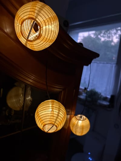 The image shows a close-up of several paper lanterns hanging from a string. The lanterns are a warm, golden color and have a striped pattern. They are illuminated from within, casting a soft glow. In the background, a window is visible, with sheer curtains partially drawn, revealing a blurry view of trees and the sky, suggesting it is either dawn or dusk. The scene appears to be indoors, near a wooden piece of furniture, possibly a cabinet or wardrobe, which has a reflective surface that mirrors one of the lanterns. The overall ambiance is cozy and atmospheric, with the dark tones of the furniture and background contrasting with the warm light of the lanterns. The location is identified as Delbrück, Germany. There are no people or animals visible in the image.