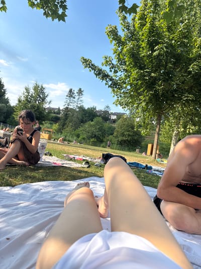 The image depicts a summer day at an outdoor gathering, likely a park or beach area, in Bürgstadt, Germany. The scene is filled with people relaxing and enjoying the warm weather. In the foreground, a person's legs are visible, covered by a white cloth, suggesting they are lying down on a blanket or towel. Next to them, another person's torso and arm are visible, also relaxed.

Further back, a woman is seated on the grass, engrossed in her phone while holding a towel. Other individuals are scattered across the grassy area, some appearing to be sunbathing or sitting on blankets. The background features lush green trees, a bright blue sky with scattered clouds, and buildings in the distance, hinting at a suburban or semi-rural environment. The overall atmosphere is one of leisure and enjoyment, characteristic of a pleasant summer outing. The time of day appears to be afternoon, given the quality of light and the activities shown. There is no visible text in the image.