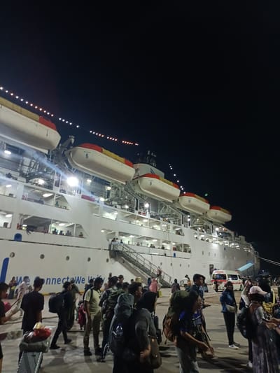 At night in Batam, Indonesia, a large white passenger ship is docked. A crowd of people, many carrying backpacks, are gathered on the pier near the ship. An ambulance is parked next to the ship. The ship has multiple levels with rows of lifeboats secured on top. The phrase "We connect We Unity" is visible on the side of the ship.