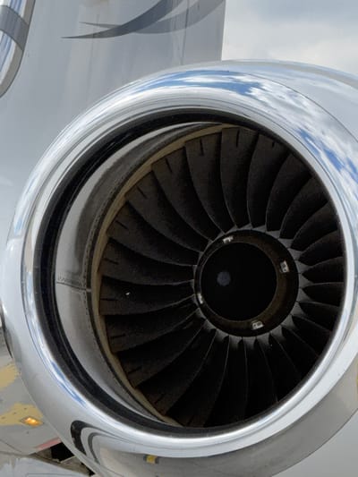 A close-up photograph captures the intake of a jet engine, showcasing its intricate fan blades and highly reflective metallic nacelle. The central focus is the dark, spiraling fan blades, clearly visible and stationary, indicating the engine is not running. The blades radiate from a dark central hub, and small, light-colored markings resembling letters like 'B' and 'E' are discernible on the inner rim of this hub, possibly indicating sensors or inspection points.

The polished, chrome-like surface of the engine nacelle is highly reflective, mirroring a bright blue sky with scattered white clouds, suggesting clear or partly cloudy daylight conditions. These reflections are distorted by the curved surface of the engine. Towards the lower right of the engine, a small, amber-colored light or sensor is visible on the exterior of the nacelle.

Part of the aircraft's light gray fuselage or wing is visible to the left and bottom left of the engine, featuring subtle, darker gray striped markings. The image gives an impression of the aircraft being parked or undergoing inspection. The scene is set in Skulte, Latvia.