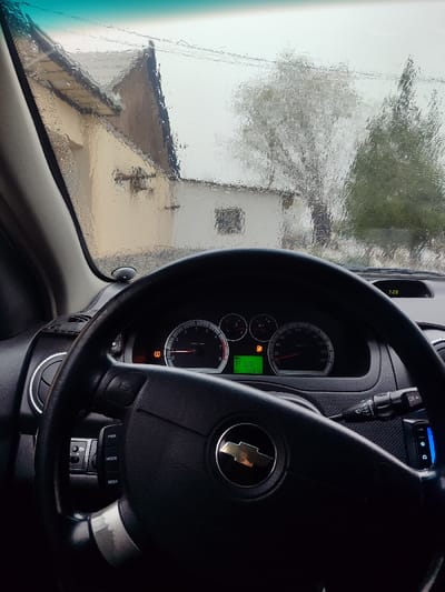 The image depicts the interior of a vehicle, specifically the dashboard and a view through a rain-streaked windshield, looking out onto a residential area in Rishtan, Uzbekistan. The foreground is dominated by a black steering wheel with a silver Chevrolet bowtie emblem in its center. The left spoke of the steering wheel contains buttons labeled "PWR," "MODE," and "SEEK." To the left of the steering wheel, a circular air vent with a chrome ring is visible.

Behind the steering wheel, the instrument cluster is visible, featuring a tachometer, speedometer, fuel gauge, and temperature gauge. Two orange warning lights are illuminated on the cluster: one resembling a tire pressure monitoring system (TPMS) warning, and another appearing to be a fuel pump icon, possibly indicating low fuel or a check engine condition. A digital display within the cluster shows "P 168.6" on a green background, with "1287" below it. A separate digital clock/temperature display on the upper right of the dashboard reads "7:28." A wiper control stalk extends from the right side of the steering column.

Through the water-laden windshield, the exterior scene appears blurred. A light-colored, possibly cream or beige, building with a dark, sloped roof is prominent on the left. Adjacent to it, a portion of a smaller white building with a dark roof and a window is visible. Beyond the structures, two trees are discernible: one with lighter, possibly bare branches, and another darker, evergreen-like tree to its right. The sky appears overcast, contributing to the diffused lighting of the scene. No individuals are visible, and the vehicle appears to be stationary.