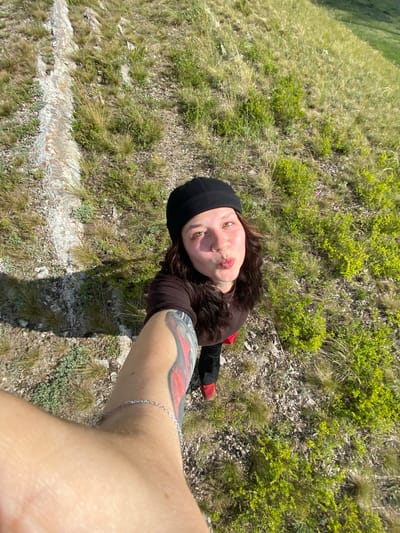 A woman is taking a selfie outdoors on a grassy hillside. She is wearing a black beanie, a dark brown t-shirt, and black pants with red shoes. She has a tattoo on her left arm and a silver anklet on her right leg. The background shows a rocky, vegetated slope. The scene is set in Andreevka, Russia.