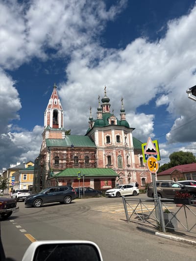 The image shows a street scene in Pereslavl-Zalessky, Russia, on a cloudy day. In the foreground, a dark-colored car is parked on the right side of the road. Further down the street, a white car is parked, and a person can be seen walking nearby. On the left side of the road, a historic brick building with green domes and white ornamentation, likely a church, is prominently featured. The building appears to be undergoing some restoration work, with scaffolding visible in parts of it. A speed limit sign displaying "50" is prominently displayed on a post, along with a triangular warning sign indicating a bumpy road. The sky is filled with dramatic, white and grey clouds against a deep blue background. The overall impression is that of a typical day in a Russian town, with a mix of modern vehicles and ancient architecture. The lighting suggests it is daytime, likely late morning or early afternoon.
