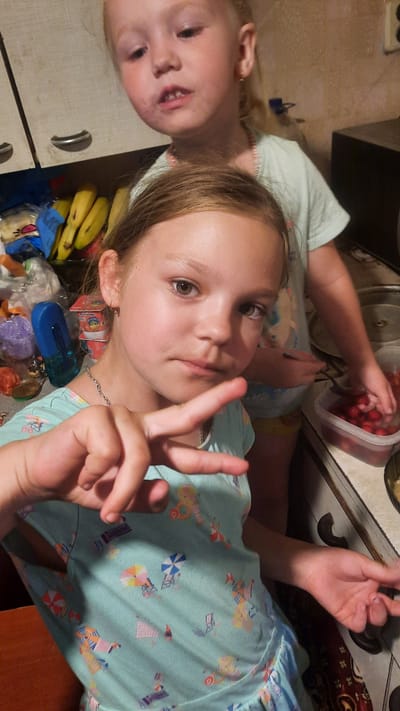 The media file shows two young girls in a kitchen. The girl in the foreground is looking towards the camera and holding up two fingers in a peace sign. She is wearing a light blue shirt with a pattern of beach scenes, including umbrellas and beach chairs. Her hair is brown and she has light-colored eyes. Behind her and slightly to the left, another young girl with blonde hair is visible. She is wearing a similar light blue shirt. The kitchen counter is visible, with a clear plastic container of red berries on it. There are also some bananas in a bunch near the bottom of the frame. The lighting suggests it is daytime. The scene appears to be in a home kitchen.