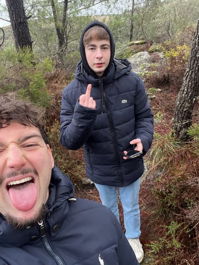 This image captures two males in a natural, overgrown outdoor environment within Ribeira de Pena, Portugal. In the foreground, an adult male with dark, wavy hair, a mustache, and a short beard, is seen in a close-up selfie orientation. He wears a dark blue puffer jacket featuring a white polo player logo and is depicted with a playful expression, his eyes squinted, mouth open wide, and tongue extended. Behind and to the right of the foreground subject stands a younger male, likely an adolescent or young adult, with light brown hair. This individual wears a dark blue hooded puffer jacket with a green crocodile logo (Lacoste), light blue jeans, and white sneakers. His right hand is raised, with the middle finger extended towards the camera, while his left hand holds a black smartphone. The surrounding environment consists of sparse trees with dark trunks, dense undergrowth of brown and green shrubs, and scattered grey rocks, consistent with a rural, vegetated landscape under an overcast sky.