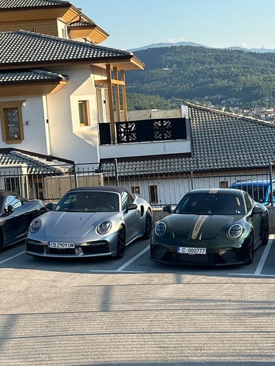 Two Porsche cars are parked on a paved surface in front of a modern building with a tiled roof. The car on the left is silver and appears to be a convertible. It has a license plate that reads "CB 2909 OM". The car on the right is dark green with gold racing stripes on the hood and roof. It has a license plate that reads "C 000777". In the background, there is a hilly landscape with trees and some buildings visible on the slopes. The sky is clear and blue. The scene is lit by natural sunlight, casting shadows on the ground. The overall impression is one of luxury and clear weather.
