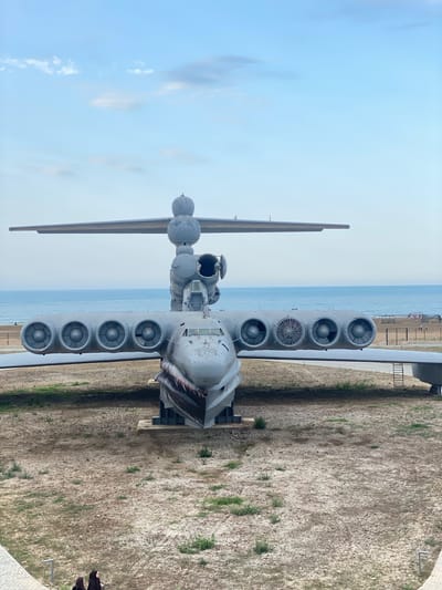 In Arablyar, Russia, a large, grey, amphibious aircraft, likely an Ekranoplan, rests on a dry, sandy area near a body of water. The aircraft exhibits signs of age and disuse, with visible weathering and what appears to be a shark-like paint scheme on its nose. It is positioned with its nose facing the viewer, its wings extended horizontally, and multiple engines mounted above the wings. The background shows a calm, blue ocean and a hazy sky with a few scattered clouds. The ground around the aircraft is barren, with sparse patches of dry grass and scattered debris. In the lower left corner of the image, two individuals, dressed in dark clothing, are partially visible, walking away from the camera. The scene appears to be an open, outdoor exhibit or a derelict aircraft site.