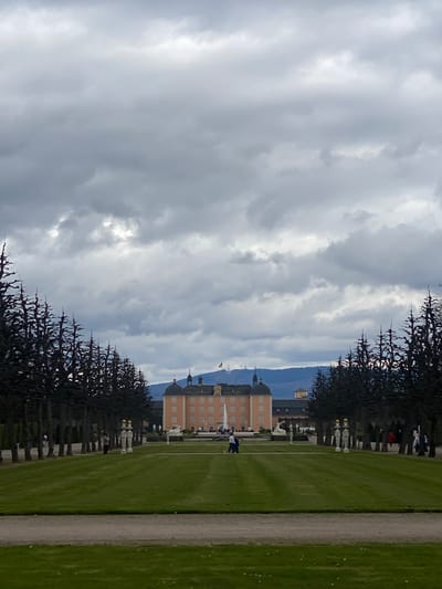 The image shows a view in the gardens of Schwetzingen Palace in Schwetzingen, Germany. The pale orange palace with its several towers is positioned in the center, receding into the distance against a background of rolling hills partially obscured by overcast skies. The foreground consists of rows of tall, pruned trees flanking both sides, leading toward the palace. A wide, graveled path runs parallel to the row of trees on the right-hand side of the image. Green lawns stretch out on either side of the path, with occasional figures of people visible. A fountain is shooting water into the air in front of the palace.