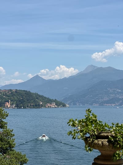 This is a landscape photograph of Lake Como in Tremezzina, Italy. The foreground features a stone planter with green foliage. In the mid-ground, a white boat is moving across the blue water, leaving a wake behind it. The background consists of a lush green hillside dotted with buildings, a prominent church tower, and a chain of misty blue mountains under a hazy sky with scattered white clouds. A small airplane is visible in the upper right corner of the sky.