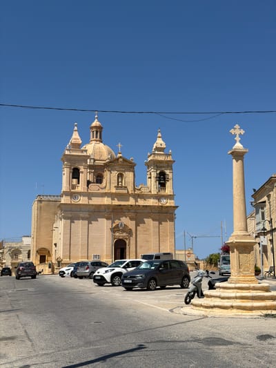 In the city of Żebbuġ, Malta, a clear, bright day unfolds in a town square. The focal point is a large, pale stone church with a dome and two bell towers, its facade adorned with architectural details. In front of the church, a row of parked cars, including a dark SUV, a white hatchback, and a grey hatchback, occupy the paved square. A stone monument, a tall, weathered column topped with a cross, stands to the right of the church. A light-colored scooter is parked near the base of the monument, its kickstand down. The sky above is a vibrant, cloudless blue, and a black wire stretches across the top of the frame. Beyond the immediate square, hints of other buildings, construction cranes, and distant foliage add to the urban landscape.