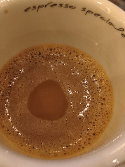 The image provides a close-up, overhead view into a white ceramic coffee cup containing a freshly made espresso. The interior wall of the cup, above the coffee line, is adorned with handwritten-style black text that spells out "espresso speciale".
The espresso itself is the central subject, featuring a thick, golden-brown crema layer that covers the entire surface. This crema exhibits a distinct circular formation: the outer ring is densely populated with numerous small bubbles, creating a textured and lighter brown appearance. Towards the center, the crema is smoother, slightly darker, and appears as a small, somewhat recessed, abstract shape, possibly resembling a teardrop or heart, where the liquid underneath is more visible. Glimmers of light reflect off the surface of the crema, adding to its visual richness and suggesting its freshness.
The cup is a simple, off-white or cream color, providing a neutral backdrop for the vibrant coffee. The lighting appears soft and warm, enhancing the inviting tones of the espresso. The photograph has a slightly grainy texture, which contributes to its overall aesthetic. No individuals are visible, and the setting is focused solely on the coffee in the cup. The image captures a serene moment, emphasizing the visual appeal and quality of the espresso.