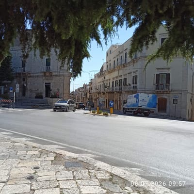 In Ruvo di Puglia, Italy, a street scene unfolds under a clear blue sky. The foreground features a cobblestone sidewalk with two circular metal manhole covers and one rectangular metal grate. A grey asphalt road with white lane markings dominates the midground, curving gently to the left. A silver minivan is turning left onto the road from the foreground. Further down the road, a dark SUV is traveling in the same direction as the minivan. On the right side of the road, a white truck with a blue and white geometric design on its side is parked. The truck has "DI TACCHIO" written on its side.
The background is lined with old buildings, characterized by their white stone facades, multiple balconies with white railings, and dark-shuttered windows. Some buildings have arched doorways and ornate architectural details. Trees with dark green foliage hang over the street from the left side of the frame, partially obscuring the view of the buildings. Traffic signs, including a blue sign with a white bird and directional arrows, are visible on the right side of the road. A traffic light and a "P" parking sign are present on the left side of the intersection. The image is time-stamped with "06.07.2026 09:57".