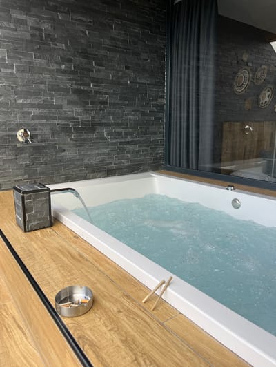 This is a medium shot image of a jacuzzi in a bathroom. The jacuzzi is filled with blue water and bubbling. On the wooden edge of the jacuzzi, there is an ashtray with cigarette butts and two unlit cigarettes. The walls of the bathroom are made of dark grey stacked stone. A chrome faucet is visible on the wall above the jacuzzi. A window is present in the background, showing a reflection of a bed and a wooden headboard. The flooring is made of wood-effect tiles.