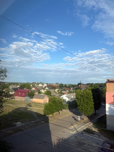 This is an eye-level, medium-distance, vertical shot of a residential area in Sosnivka, Ukraine, under a bright blue sky dotted with scattered white clouds. The scene depicts a mix of single-family homes with pitched roofs in various colors, including red, grey, and brown, interspersed with lush green trees. A paved road with visible tire marks runs through the middle ground, casting long shadows. Power lines crisscross the frame, originating from a multi-story brick building on the right side of the image, which features a pink and white facade. The foreground is dominated by the shadows of these power lines and the building. A dark-colored car is partially visible in the lower right corner. The overall environment suggests a tranquil, suburban setting during daylight hours.