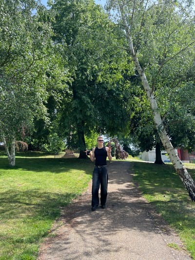 A figure stands on a gravel path in Christchurch Park, Ipswich, UK. The park is characterized by mature trees providing substantial shade. The figure, presumably male, is attired in dark trousers and a dark tank top. He wears sunglasses and a hat. The figure appears to be holding a phone. A birch tree stretches across the scene. The lighting indicates daylight. The scene includes a path and grassy areas. There is a fence on the park boundary.