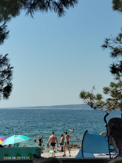 The image shows a sunny day at a pebble beach on the island of Krk, Croatia. The water is a clear blue and there are several people swimming and relaxing. In the foreground, there are two blue beach tents and a stroller. The beach is lined with rocks and trees. In the background, there is a distant coastline. The overall scene depicts a peaceful day at the beach.