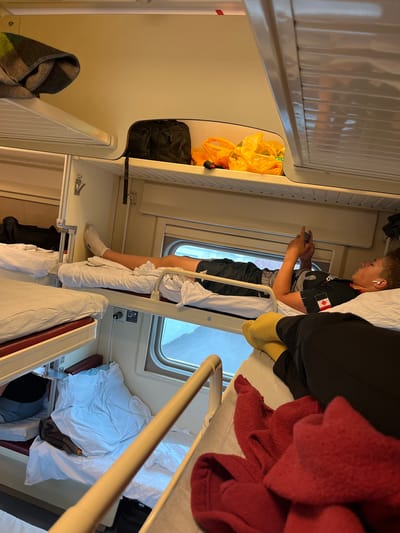 The image depicts the interior of a train compartment, specifically a sleeper carriage, with multiple bunks visible. In the upper bunk on the right, a young person is lying down, looking at a smartphone. They are wearing a black t-shirt with a red and white emblem on the left side of the chest and white earbuds. Below them, a pair of yellow socks is visible on someone else's feet. On the left side of the compartment, a blanket is neatly folded on an upper bunk, and a black bag rests on a shelf. The overall environment is characterized by the typical setup of a train sleeping compartment, with beige walls, white linens, and metal railings. The train appears to be in motion, as suggested by the window view which shows a blurred outdoor scene. The location is identified as the city of Vyatskiye Polyany, Russia.