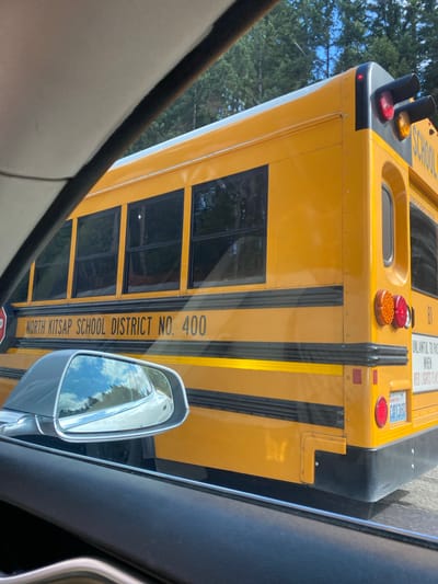 An exterior, eye-level, medium shot captures the rear section of a yellow school bus, identified as belonging to the "NORTH KITSAP SCHOOL DISTRICT NO. 400," as it travels on a road. The bus is the primary subject, with its distinctive orange and red flashing lights and brake lights visible at the top rear. The side of the bus is emblazoned with the district name and number. A white sign on the rear of the bus reads "UNLAWFUL TO PASS WHEN RED LIGHTS FLASH." A license plate with the identifier "C0138D" and "WASHINGTON" is visible. A silver car's side mirror is reflected in the image, showing the sky and trees. The background consists of a dense forest with tall evergreen trees and a bright blue sky with scattered clouds. The bus appears to be in motion, suggesting it is on a route through a semi-rural or suburban area of Port Orchard, Washington, United States of America.