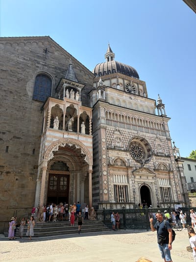 The image shows the Basilica di Santa Maria Maggiore in Bergamo, Italy, on a clear, sunny day. A crowd of tourists is gathered on the steps leading up to the church entrance. The basilica's facade is elaborately decorated with marble and stone, featuring statues, intricate carvings, and a large rose window. The sky is a bright blue.