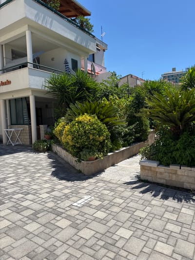 The image depicts a building entrance and courtyard area in Makarska, Croatia. The building is white with a covered balcony area supported by pillars. A sign with the word "Vanko" is visible on the building's exterior. The courtyard is paved with interlocking rectangular and square grey stones arranged in a geometric pattern. To the right of the building entrance, a landscaped garden features various green plants, including palm-like foliage and a dense, rounded bush with yellow-green leaves. The garden is bordered by a stone wall. The sky is clear and blue, indicating a sunny day. The scene appears to be a residential or commercial property with an emphasis on outdoor landscaping and a well-maintained courtyard.