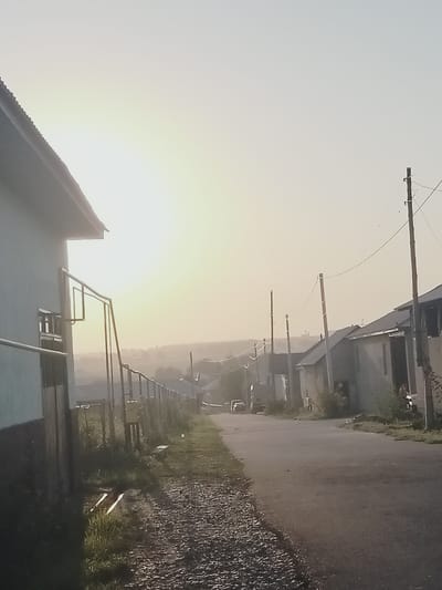 The image depicts a street scene in the city of Көлкент, Kazakhstan, bathed in the soft, hazy light of a low sun. On the left, a light-colored building with a dark, sloped roof partially frames the view. A metal fence runs along the side of the road, separating the property from the street. The road itself is paved and curves gently into the distance. Several utility poles with wires crisscross the sky. On the right side of the road, houses with dark roofs are visible, some appearing to be under construction or in a state of disrepair. The landscape in the background is indistinct, appearing as a hazy, distant horizon. There are a few vehicles present, including a white truck and a car, parked or driving on the road. The ground beside the road is covered with gravel and grass. The overall atmosphere is quiet and somewhat desolate, suggesting a rural or semi-rural setting within the city.