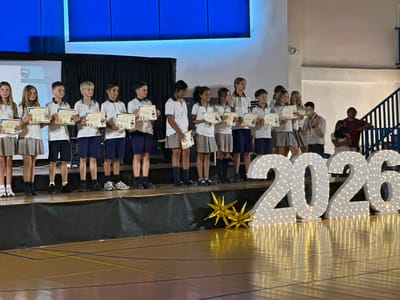 A group of children and adults are standing on a stage. The children are lined up and holding certificates. The adults are standing behind them or to the side. There are large illuminated numbers "2026" in the foreground. The scene appears to be an awards ceremony or graduation event. The children are dressed in school uniforms. The background shows a stage with a black curtain and a projector screen. The setting appears to be indoors, possibly a school gymnasium or auditorium. The lighting suggests it is evening or indoors.
