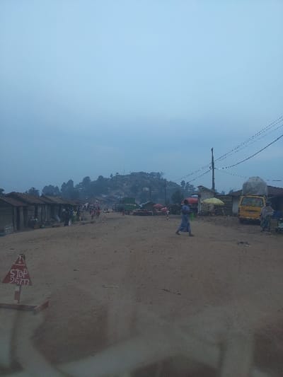 The image depicts an unpaved main thoroughfare in Kirumba, Democratic Republic of the Congo, under an overcast, light blue-grey sky. A wide dirt road occupies the foreground and midground, extending into a hazy distance.
On the lower left, a red and white triangular sign on a wooden stand displays "STOP 25m". Along the left side of the road, a continuous row of single-story structures, constructed from wood paneling or corrugated metal, with matching roofs, appears to function as market stalls or shops. Several individuals are visible standing or moving near these structures.
In the center midground, a lone individual dressed in a light blue full-length garment walks from right to left across the dirt road, carrying a green bucket or bag in their right hand.
On the right side of the road, a yellow utility van is parked, with a substantial, white tarp-covered object secured to its roof. Adjacent to the van, additional market setups are visible, characterized by red and yellow parasols. A utility pole with overhead power lines extends vertically from the right midground. Further down the road, numerous other pedestrians are observed.
The background features vegetated hills with scattered residential or commercial structures, partially obscured by atmospheric haze.