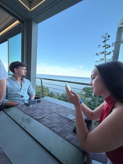 Two individuals are seated at a table on a balcony overlooking the sea in Santa Maria Navarrese, Italy. The man, on the left, is wearing a light blue striped button-down shirt and looking out at the water. He has dark curly hair and is wearing a watch on his left wrist. In front of him, there is a glass and cutlery. The woman, on the right, is wearing a red top and is looking down at her phone. She has long dark hair and is wearing a blue wristband. The table is covered with a patterned cloth. Beyond the balcony, the sea stretches to the horizon under a clear blue sky, with some greenery visible on the lower slopes. The architecture of the building with modern lighting fixtures suggests a contemporary establishment.
