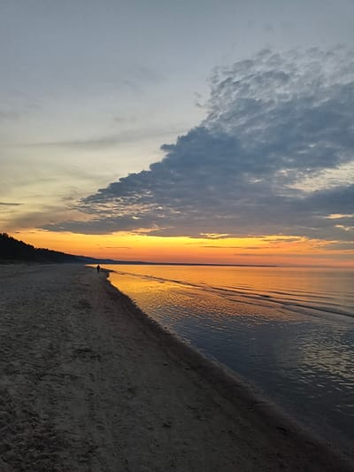 A solitary figure walks along a sandy beach at dusk in Apšuciems, Latvia. The sky is a canvas of transitioning colors, with soft grays and whites fading into vibrant oranges and yellows near the horizon. Dark, textured clouds drift across the upper portion of the sky, contrasting with the smoother, lighter clouds nearer the sunset. The calm sea reflects the warm hues of the sky, creating a shimmering, golden path across its surface. Gentle waves lap at the shore, leaving patterns of foam on the wet sand. The beach itself is a dark expanse of sand, marked with footprints, leading towards a line of dark trees on the left side of the image. The scene evokes a sense of peaceful solitude and the beauty of nature at the end of the day. The time of day is clearly sunset. The weather appears to be calm with light clouds. There is no visible text in the image.