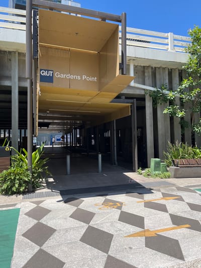 This image captures an outdoor pathway and the entrance to a building, likely part of a university campus in Brisbane City, Australia.

The main subject is a large, golden-colored sign prominently displaying the text "QUT Gardens Point" in white lettering, indicating the Queensland University of Technology's Gardens Point campus. This sign is situated on the left side of the frame, spanning a covered walkway or underpass.

To the left of the sign, a concrete staircase with white railings ascends, integrated into the building structure and partially adorned with green foliage and trees. The pathway itself leads into a shadowed area, revealing further structural pillars and overhead elements, and a smaller "QUT" sign visible deeper within the passage.

The ground on the right features a distinctive checkerboard pattern of alternating light grey and dark grey square tiles. Painted on this footpath are yellow directional arrows and a yellow bicycle symbol, suggesting a designated shared path for pedestrians and cyclists. A thin green strip along the very edge of the footpath also indicates a cycling lane. Further down the path, a green recycling bin and some potted plants are visible.

The scene is bathed in bright daylight under a clear blue sky, casting distinct shadows across the patterned footpath, suggesting a sunny day. There are no people visible, presenting a quiet view of the campus grounds.