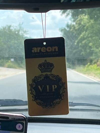 A close-up shot shows an Areon "VIP EXCLUSIVE" air freshener hanging from a string inside a vehicle, with the windshield and dashboard visible. The air freshener is gold and black, featuring a crown and ornate scrollwork. Outside, a road lined with trees and a cloudy sky are visible through the windshield. The image is taken from the interior of a car, likely on a road in or near Asparuhovo, Bulgaria.