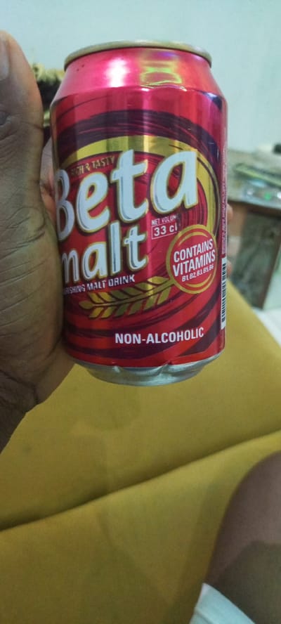 A close-up, eye-level shot shows a person's hand holding a red and gold can of Beta Malt, a non-alcoholic malt drink, in Omagwa, Nigeria. The can has prominent white lettering that reads "Beta malt" and "NON-ALCOHOLIC," along with text indicating it "CONTAINS VITAMINS B1, B2, B3, B5, B6" and a net volume of "33 cl." The background is blurred, showing a yellow surface that appears to be a cloth or cushion, and a hint of a wall in the upper left. The lighting suggests an indoor setting, possibly during daylight hours. The image captures a casual moment, with the can being the central subject.