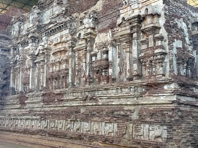 The image shows a detailed, close-up view of a weathered brick structure, specifically the exterior wall of the Thuparama Vatadage in Polonnaruwa, Sri Lanka. The structure exhibits intricate architectural details with repetitive columns, moldings, and layered brickwork. The colors are muted, primarily consisting of browns, grays, and whites, suggesting age and exposure to the elements. The wall also features a lower level with carved figures or motifs in relief, further displaying the ornate nature of the construction. The overall condition of the brickwork appears worn and deteriorated in places, hinting at the historical significance and antiquity of the site.