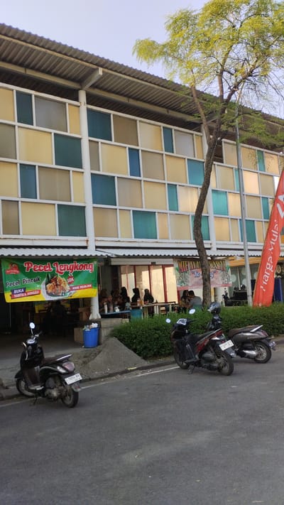The image shows a street scene in Trosobo, Indonesia, outside a building with a multi-colored facade and a corrugated metal roof. A prominent green banner advertising "Pecel Lengkong" is displayed in front of the building, alongside smaller banners for "MIE AYAM" and "HARGA MULAI". People are seated at tables outside the building, indicating an outdoor dining area. Several motorcycles are parked along the side of the road in front of the building. A large tree with light green foliage grows to the right of the building, casting a shadow. The sky is clear and bright, suggesting daytime. The overall environment appears to be a commercial area with food vendors.