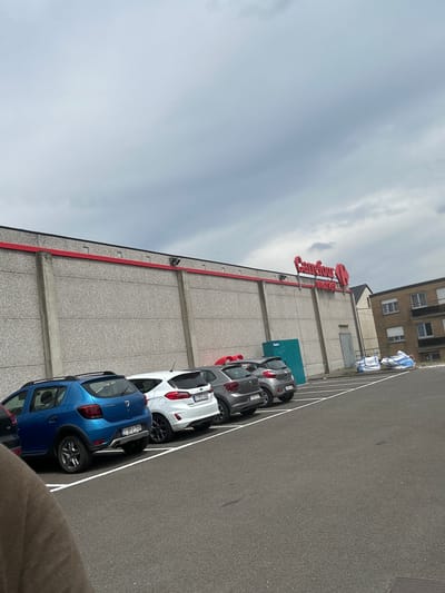 The image shows a parking lot in front of a Carrefour Market store. The store has a concrete facade with a red trim along the top. The Carrefour Market logo is visible in red letters on the building. Several cars are parked in the lot, including a blue Dacia Sandero, a white Ford Fiesta, and two gray Volkswagen Polo. The sky is overcast. The location is confirmed to be in Herstal, Belgium, based on the license plates and architectural style. There are no people visible in the image.