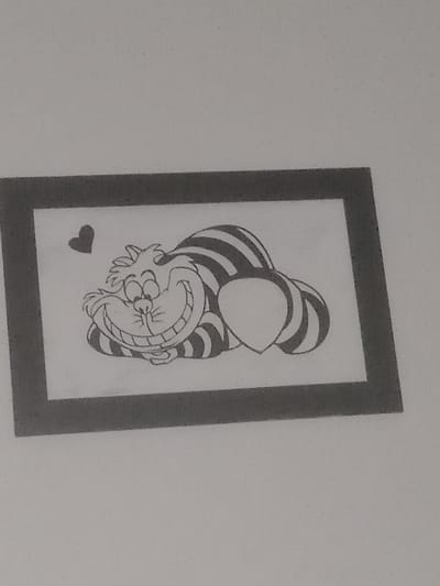 A photograph of a grayscale drawing of the Cheshire Cat from "Alice in Wonderland" inside a black rectangular frame. The Cheshire Cat is depicted upside down with his signature wide grin and striped tail. A small black heart is located at the bottom of the drawing, slightly to the left. The location is Villa Ballester Noroeste, Argentina.