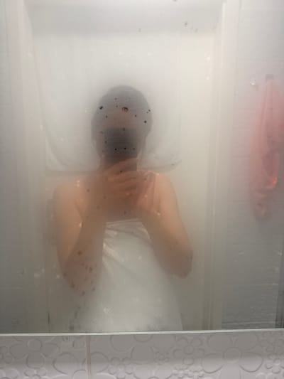 The image shows a person lying in a bathtub filled with water, as seen through a steamy glass shower door. The person has a dark-colored head covered with what appears to be black dots. The person's arms are crossed over their chest. A pink object is floating near the top of the tub. The exact location in Kranj, Slovenia is not ascertainable from the image. The bathroom has textured wall tiles with a floral pattern on the right side of the image.