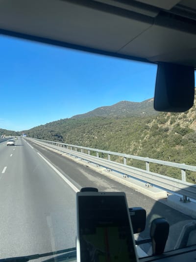 The image displays a forward-facing view from inside a vehicle traveling on a multi-lane highway in Le Perthus, France. A section of the vehicle's dark gray dashboard is visible at the bottom. Mounted centrally on the dashboard is a dark smartphone or tablet, its screen displaying a navigation application with green and red route lines. Above the device, the vehicle's clear windshield offers an unobstructed view of the highway and surrounding landscape. A dark rearview mirror housing is partially visible in the upper right corner, attached to the vehicle's ceiling lining.
The highway, constructed of asphalt, extends directly forward and slightly to the left, marked with clear white solid and dashed lane lines. Several distant vehicles, including a distinct white car, are visible in the left lane, heading away from the viewer. Along the right side of the highway, a metal guardrail runs parallel to the direction of travel, supported by concrete barriers. Beyond this guardrail, a steep mountainside covered in dense green vegetation descends, while a higher, rockier mountain peak is visible further in the background. The sky above is clear and bright blue, indicating sunny weather conditions.