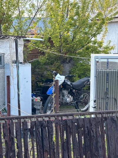 The image, presented in a vertical orientation, captures a cluttered outdoor space. A heavily rusted, dark brown metal fence or gate extends vertically along the right side of the frame, its horizontal slats showing peeling paint and corrosion. Peering through the fence and gaps in the foliage, various building structures with brickwork and corrugated metal roofs are visible in the background.

Dominating the mid-ground is a dirt bike-style motorcycle, primarily blue and black, with its engine and parts clearly visible. A black bag rests on its seat. Above the motorcycle, a white appliance, likely the back of a refrigerator or freezer, features prominent black condenser coils. Repeated text, "G-PENTANE", is visible on blue labels on this appliance. Another white appliance is partially seen below the motorcycle and to the left of the fence. White wires or ropes are strung across the lower section of the image, intertwining with some rusty metal bars.

The left and background areas are filled with abundant green foliage from large trees. The scene is illuminated by natural daylight, suggesting clear weather. The setting appears to be a utilitarian backyard or storage area in Aksay, Kazakhstan, characterized by a mix of natural elements and discarded or stored objects. Other visible text includes "X401" and "YADI" on different parts of the motorcycle. No discernible activity is taking place.