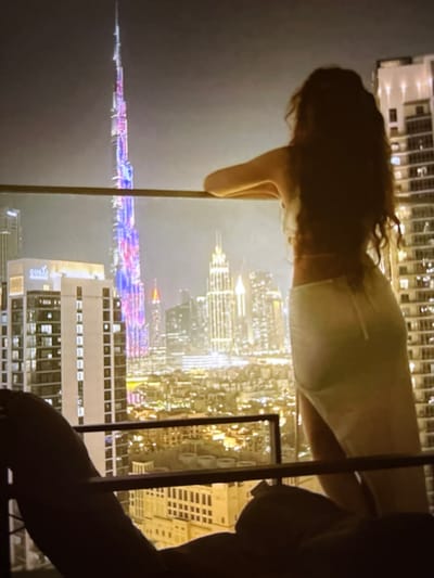 A woman stands on a balcony overlooking a city skyline at night. She has long, dark, wavy hair and is wearing a light-colored, possibly white, top and skirt. The city skyline is illuminated with many lights, and a tall, slender skyscraper is visible in the background, lit with colorful lights. The woman is leaning forward, with her back to the camera, and her arms resting on the balcony railing. The lower portion of the image shows a person lying down, with their arm resting on the railing. The overall scene suggests a view from a high vantage point in a modern city at night. The location is not Pori, Finland.