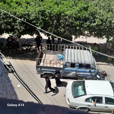 In Deir el-Balah, Palestinian Territories, a street scene unfolds under a canopy of lush green trees. A light blue pickup truck, its bed laden with what appears to be bundled fabric and boxes, is parked on the right side of the frame. Its rear is partially obscured by a white sedan, which has its passenger side door open and an arm extending out. A man in a black shirt with white detailing, wearing a medical cast on his left arm, stands near the rear of the pickup truck, looking towards the sedan.

To the left of the pickup truck, a man dressed entirely in black, including a baseball cap, walks away from the camera. He is followed closely by a woman wearing a black hijab and abaya.

Further in the background, beneath the trees, several people are gathered around what appear to be market stalls or seating areas. The ground is a mix of paved and unpaved surfaces, with scattered debris. Overhead power lines crisscross the upper portion of the image, casting shadows. The overall impression is one of a busy, sun-drenched urban environment.