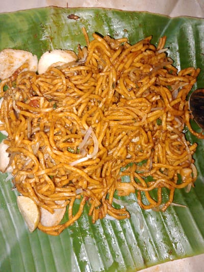 A close-up, top-down shot shows a portion of a banana leaf serving as a plate, presenting a pile of fried noodles. The noodles are thin, golden-brown, and coated in a savory sauce, interspersed with slivers of white onion and small, darker ingredients. To the left of the noodles, three pieces of a circular, white, fried item are visible, along with a wedge of lemon. The banana leaf has a vibrant green color with prominent veins, appearing slightly wet. A portion of a metallic spoon is visible on the right edge of the frame. The scene is likely from a street food stall or a local eatery in Rayeukpayaitek, Indonesia, where banana leaves are commonly used for serving food.