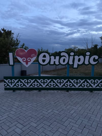 The image displays an "I Love" sign in the city of Өндіріс, Kazakhstan. The sign features the letter "I" on the left, followed by a red heart with white embellishments and the text "2005 ТУЛЕКТЕРГЕ" inside. To the right of the heart is the word "Өндіріс" in large, white, stylized letters against a green background. The sign is supported by teal-colored poles. Below the sign is a decorative green border with white traditional Kazakh patterns. The ground in front of the sign is paved with gray interlocking bricks laid in a herringbone pattern. In the background, a light-colored fence is visible, beyond which are trees and dry grass. The sky is overcast with gray clouds. There are no people visible in the image. A dark trash bin is positioned behind the fence, to the left of the sign.