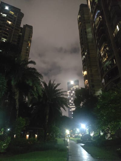 The image depicts a nighttime scene in Nanhai District, China, featuring a park with prominent residential buildings and a distant skyscraper. In the foreground, a paved walkway curves through a dimly lit park area with lush green grass and trees. Several palm trees are visible, contributing to a tropical feel. Scattered lighting fixtures illuminate parts of the park.
Dominating the background are several high-rise residential buildings, characterized by their multiple floors and numerous windows, many of which are lit, indicating occupancy. To the left, a tall, angular residential building is angled towards the sky. To the right, a curved residential building with balconies is also visible. In the mid-ground, a modern, illuminated skyscraper with a distinct facade stands out against the cloudy sky. Two figures, appearing as silhouetted individuals, are walking along the park's pathway. One is slightly ahead of the other, and they are moving away from the viewer. The overall atmosphere is tranquil, with the artificial lights of the city contrasting with the dark, natural elements of the park.
