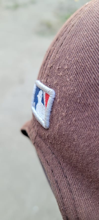 This media file shows a close-up, angled shot of a brown baseball cap. The cap is made of a textured fabric with a subtle ribbed pattern. There is an embroidered patch on the side of the cap. The patch is white with a blue border. Inside the blue border, there is a design with red, white, and blue elements, appearing to be an abstract logo or emblem. The background is out of focus, showing a light-colored, textured surface that could be concrete or pavement, and hints of green foliage suggesting an outdoor setting. The lighting appears natural, indicating it might be daytime. There is no visible text in the image. The image is focused on the texture of the cap and the detail of the embroidered patch.