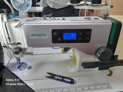 This is a close-up, eye-level, indoor photograph of a sewing machine in what appears to be a workshop or tailoring establishment. The machine is white with a silver control panel featuring the brand name "ZOJE" and a digital display showing a "P" and "S" button, along with navigation arrows and a blue screen with symbols. A small blue fan with a cartoon character is attached to the top left of the sewing machine. There are spools of black thread visible in the background. A pair of blue thread snippers lies on the table in front of the machine. A measuring tape with centimeter and inch markings is also present on the table. The image also includes a timestamp and model name overlay in the bottom left corner, indicating "Galaxy A12" and "29 июня 2026 г. 10:49". The overall scene is well-lit and focused on the sewing machine and its immediate surroundings.