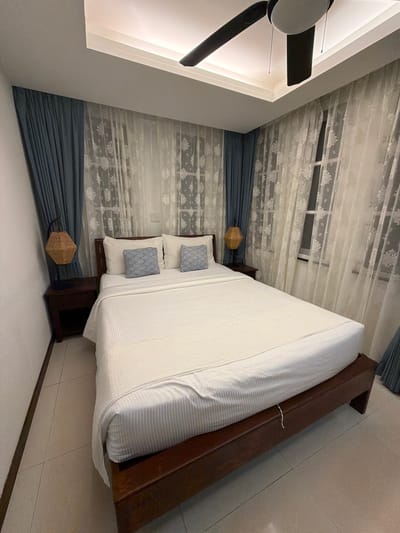 This is a forensic photograph of a well-lit bedroom in Ao Phai, Thailand. The central subject is a queen-sized or king-sized bed with a dark brown wooden frame and a matching headboard. The bed is furnished with white, horizontally striped bedding, including a fitted sheet, a flat sheet, and a bedspread. Two large white pillows are positioned against the headboard, flanked by two smaller decorative pillows featuring a blue and white wavy pattern.
To the left and right of the bed are identical dark brown wooden nightstands, each equipped with a single drawer and an open lower shelf. Suspended above each nightstand is a woven hexagonal-shaped lamp shade emitting a warm light, affixed to the wall with a dark metal fixture.
The wall behind the bed is dominated by windows covered with two layers of curtains. The inner layer consists of sheer white fabric with a floral or snowflake-like pattern. The outer layer comprises solid blue-grey drapes, which are pulled back to frame the sheer curtains. A white electrical outlet or light switch panel is visible on the wall to the right of the bed, between two window sections.
The ceiling is white and features recessed cove lighting that emits a warm, indirect glow around the room's perimeter. A black and white three-bladed ceiling fan is mounted centrally on the ceiling, with pull chains visible. The floor is covered with light-colored square tiles. The walls are painted white. No individuals or animals are present in the scene.