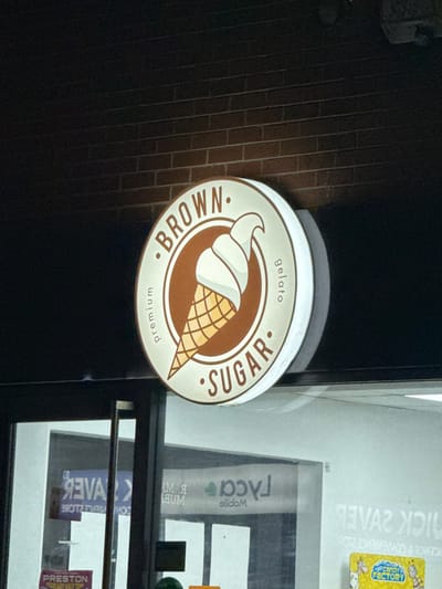 The image displays an illuminated, circular sign for "Brown Sugar premium gelato" mounted on a dark brown brick wall. The sign features a white outer ring, a central brown circle, and a graphic of a white soft-serve ice cream cone with a light brown waffle cone pattern. Below the sign, a glass storefront reflects reversed text, including "Quick Saver" (a convenience store), "Mobile," and a "Candy Factory" sticker. A "PRESTON" sticker is partially visible on the bottom left of the reflected surface. The scene appears to be at night or in low light conditions, located in the city of Preston, United Kingdom. No individuals or active interactions are present.