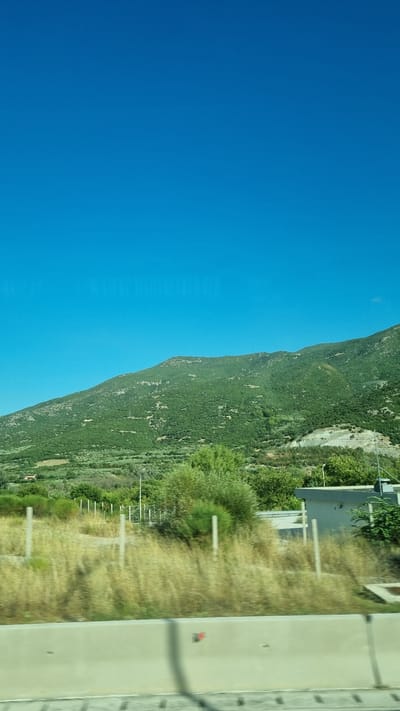 The image captures a natural landscape under a clear, vibrant blue sky. In the background, a lush green mountain slopes upwards, covered in dense vegetation. The lower portion of the image shows a dry, grassy field with scattered bushes and a fence line. To the right, a small, modern-looking building with a grey facade is partially visible, situated behind the fence. A concrete barrier runs along the bottom of the frame, suggesting a roadside perspective. The scene is set in the Municipal Unit of Ampelakia, Greece.